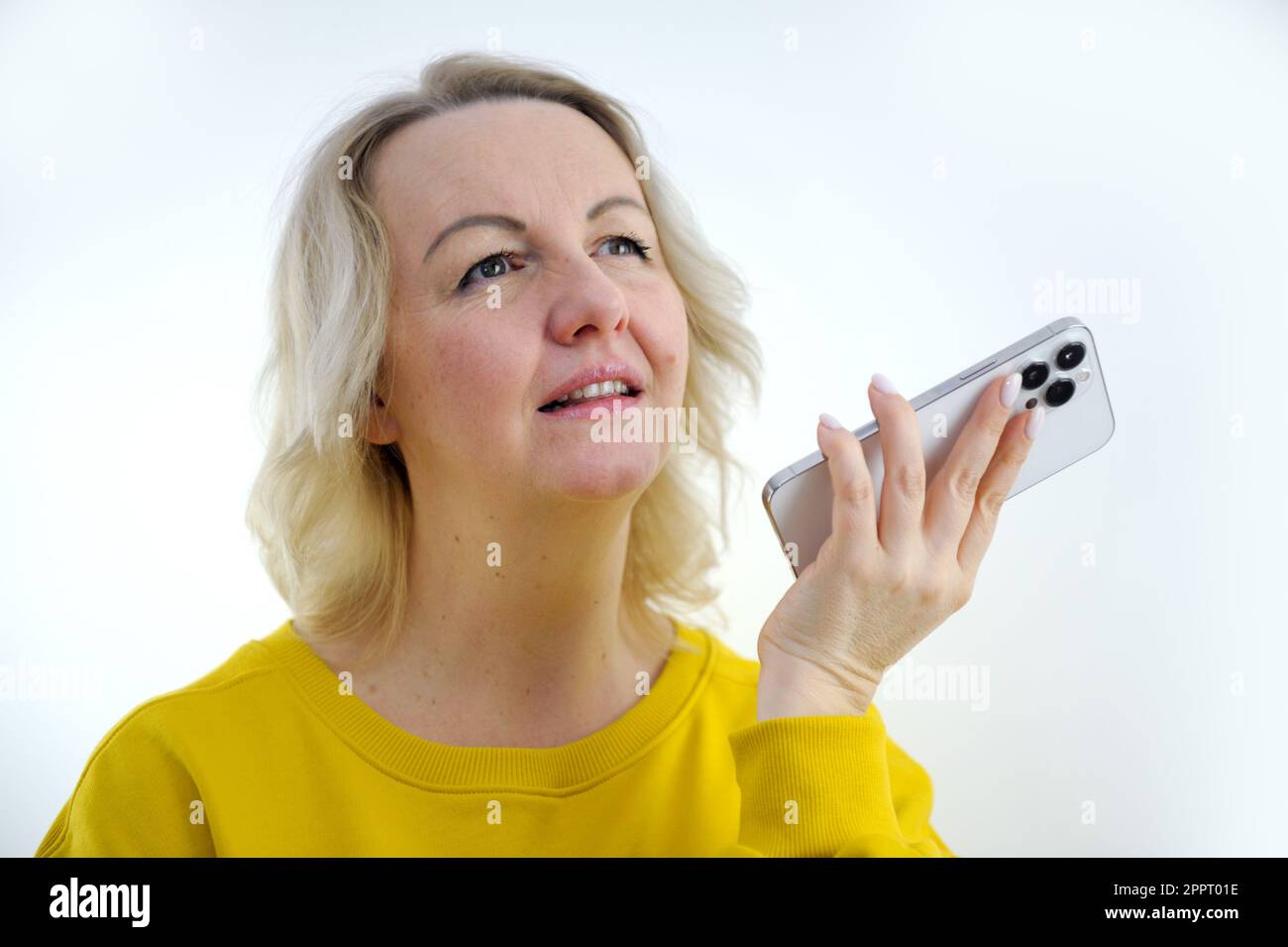 Eine Erwachsene blonde Frau, die aufmerksam nach oben schaut, hält ein Telefon mit drei Kameras in ihren Händen auf weißem Hintergrund, Gedankenzweifeln, Problemlösung, Auswahl, gute Anrufpläne für die Zukunft Stockfoto