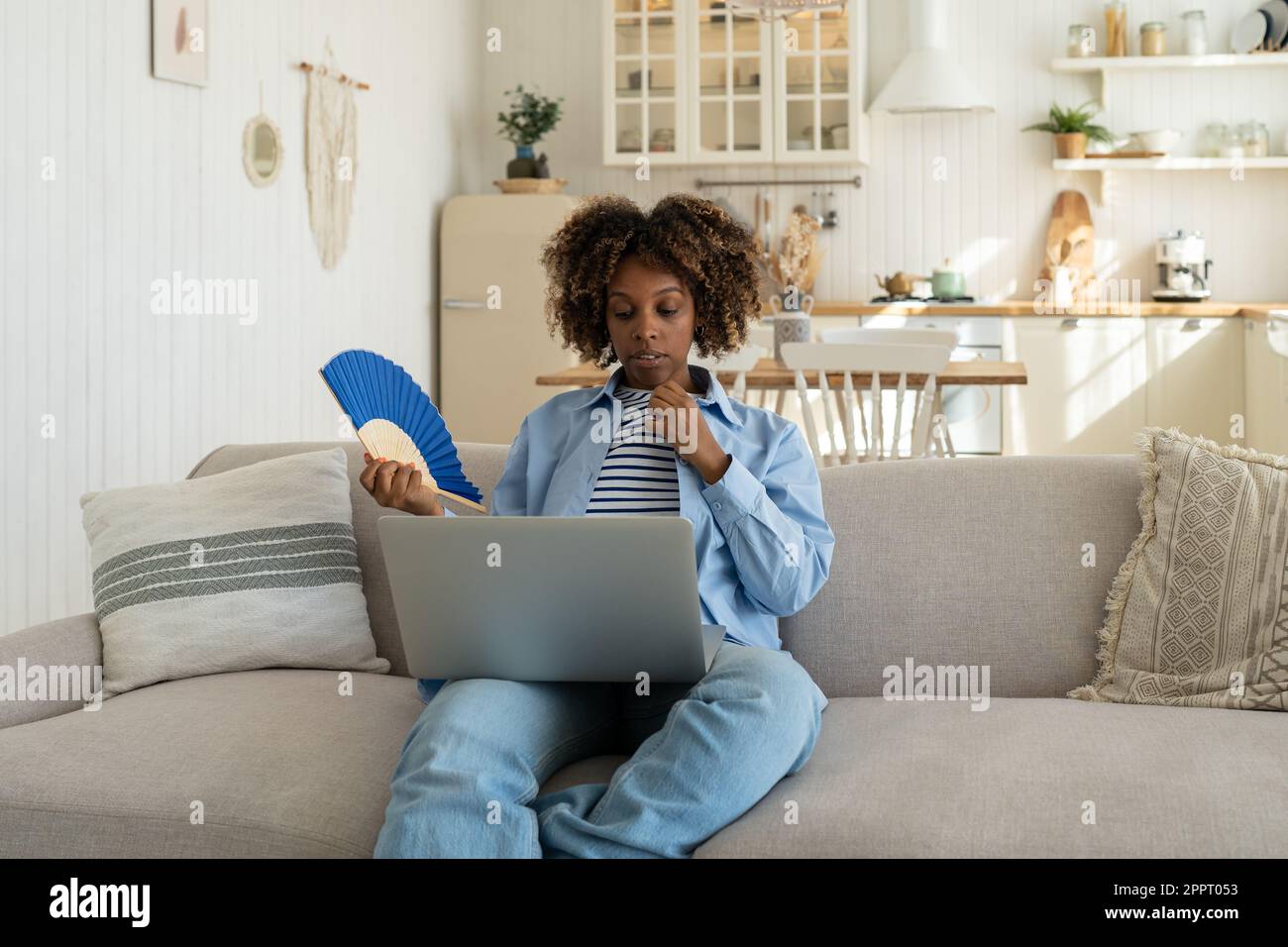 Müde, erschöpfte afroamerikanische Frau, die mit einem Ventilator wedelt und zu Hause mit einem Laptop arbeitet. Stockfoto