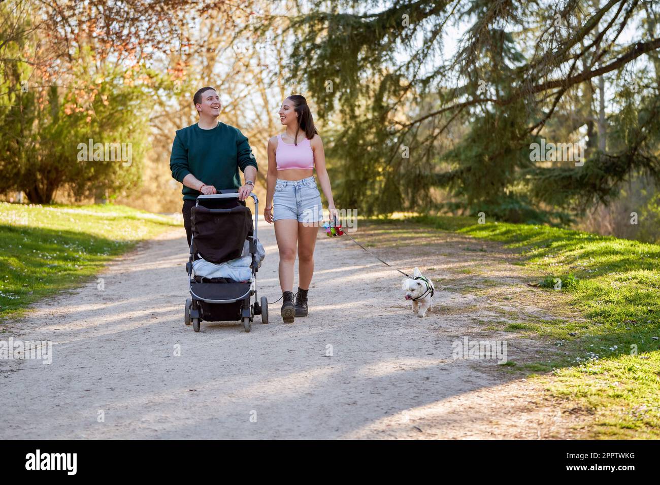 Ein junges Paar macht an einem sonnigen Sommertag einen gemütlichen Spaziergang durch den Park, schiebt sein Baby in einem Kinderwagen und begleitet von seinem treuen Hund Stockfoto