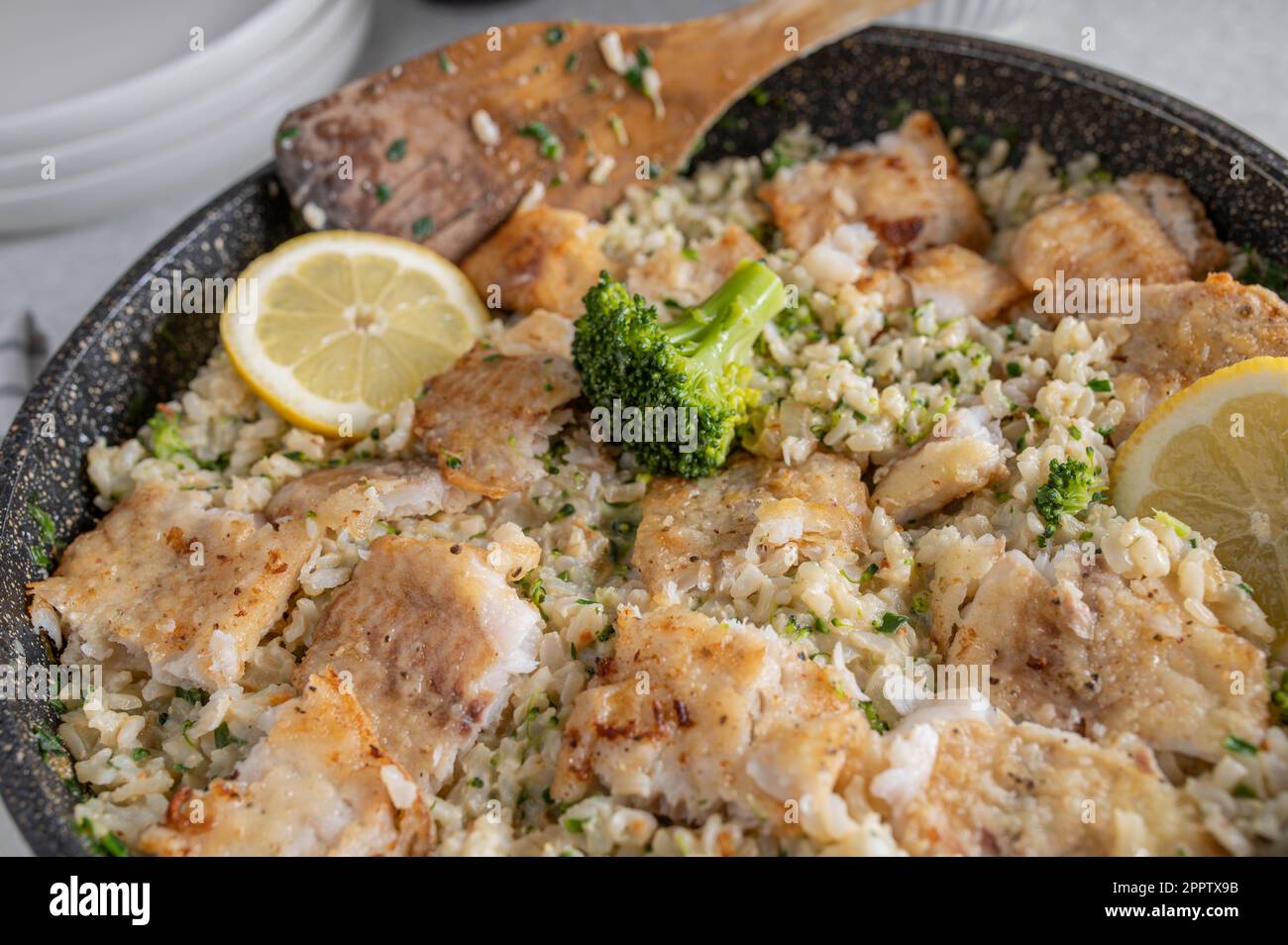Gebratenes Fischfilet in der Pfanne mit braunem Reis, Brokkoli und Schnittlauch in einer Bratpfanne auf dem Küchentisch Stockfoto