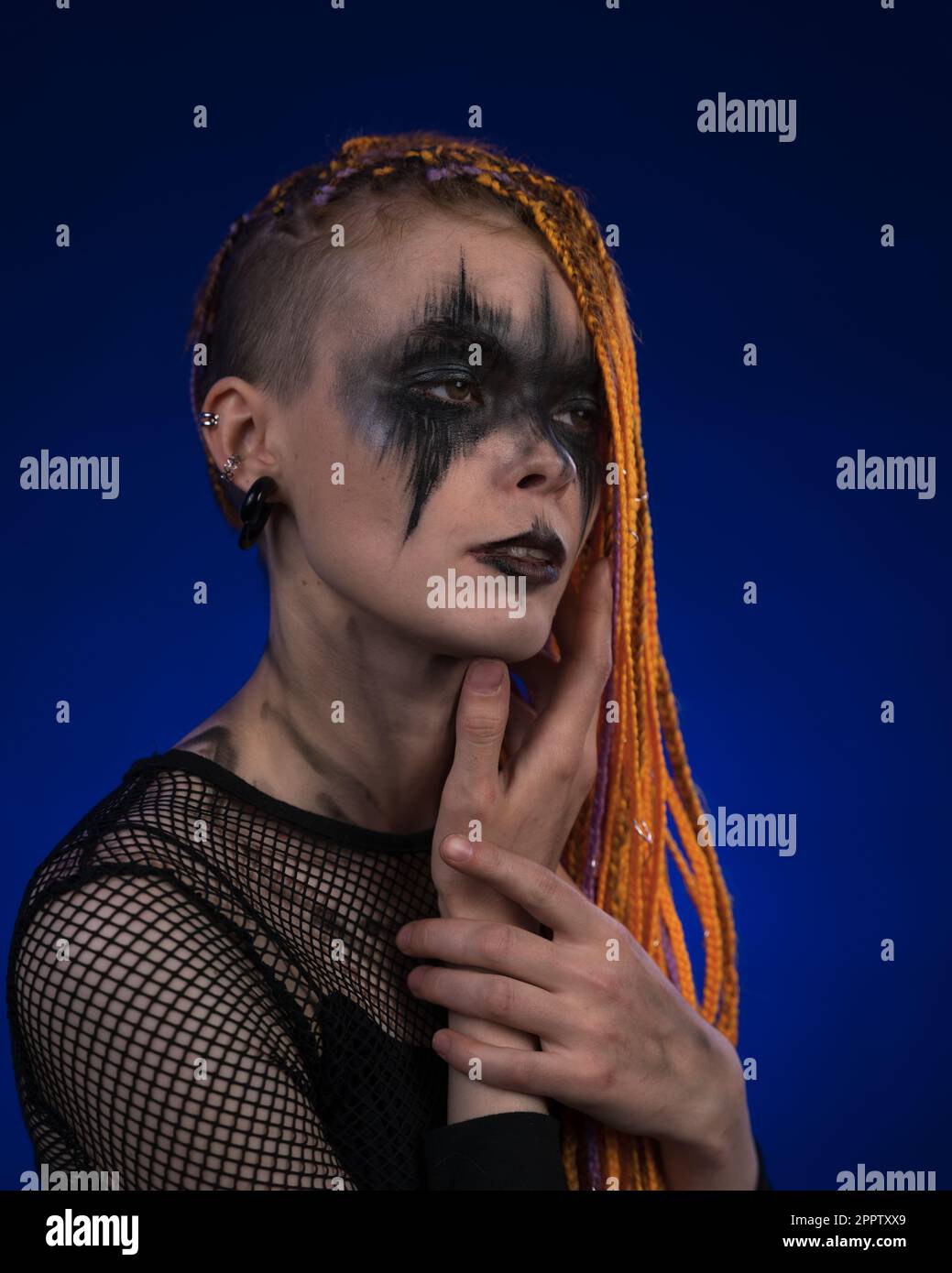 Filmporträt einer wunderschönen jungen Frau mit orangefarbenem Dreadlocks-Haarschnitt und grauenschwarzem Bühnenschmink auf dem Gesicht. Studio auf Blau aufgenommen Stockfoto
