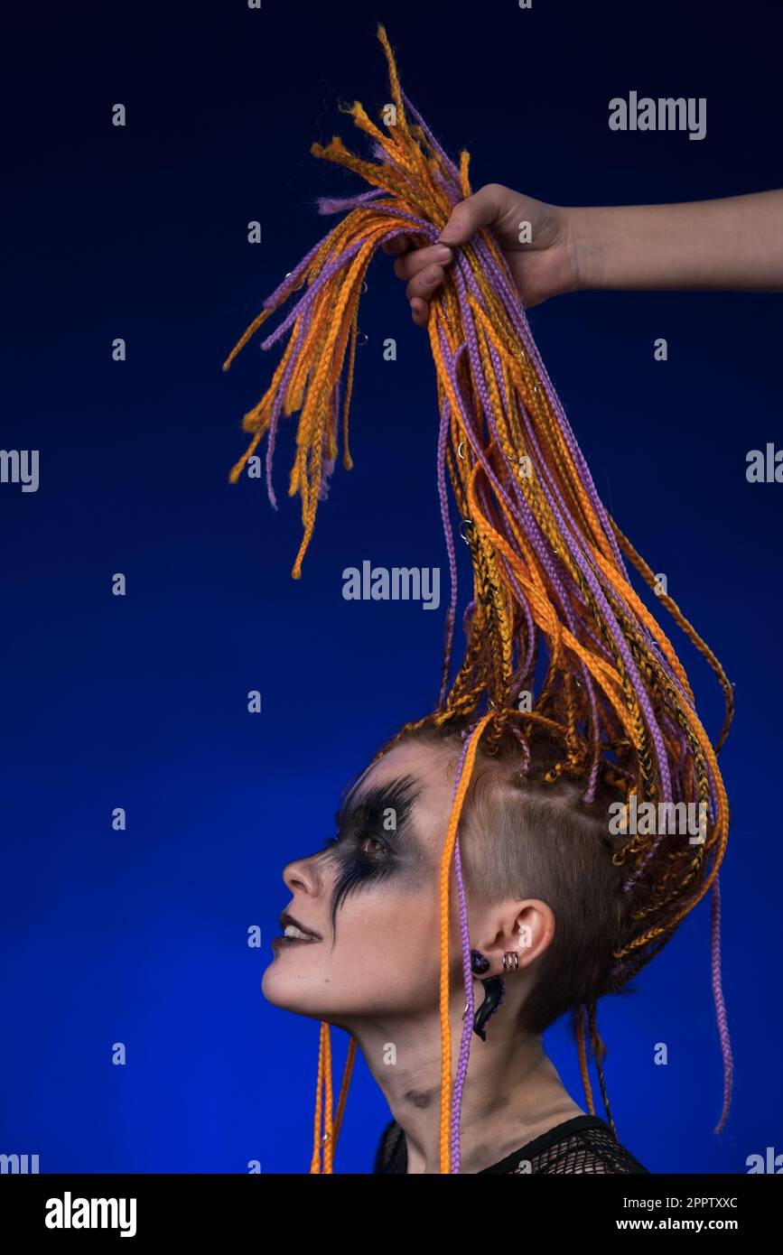 Feines Kunstporträt einer wunderschönen jungen Frau mit orangefarbenem Dreadlocks-Haarschnitt und grauenschwarzem Bühnenschmink auf dem Gesicht. Studio mit seitlichem Blick Stockfoto