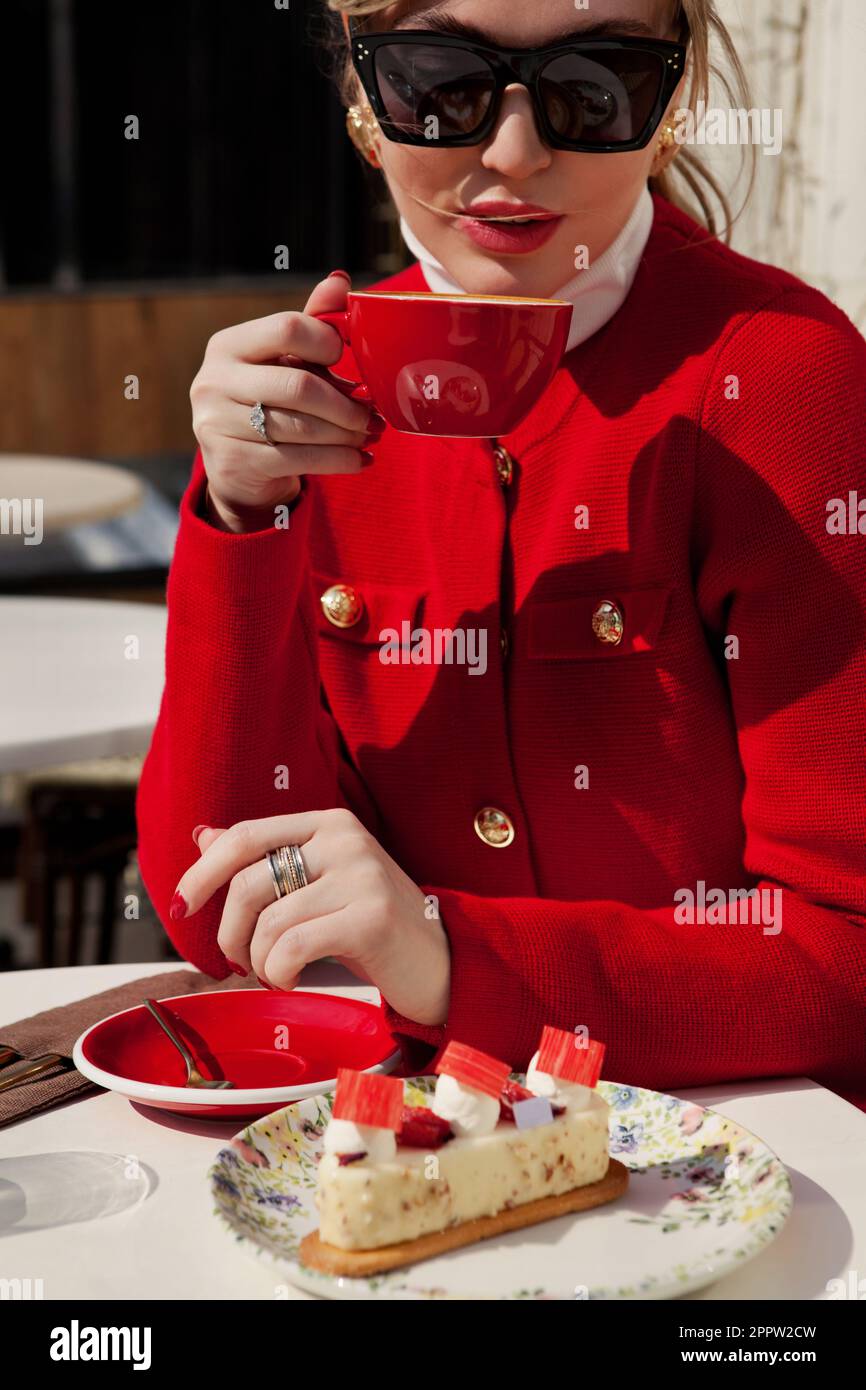 Portrait stilvolle Frau in Sonnenbrille und rotem Blazer trinkt Cappuccino-Kaffee und isst Kuchen im Straßencafé. Beende das elegante Mädchen mit einer Tasse Kaffee Stockfoto