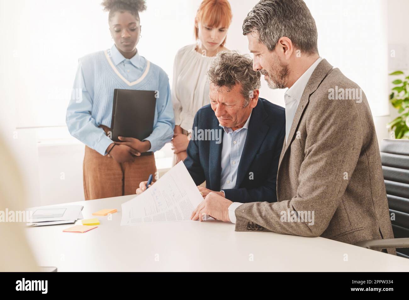 Ein Geschäftsmann hat einen Vertrag für einen älteren Menschen, der an einem Schreibtisch in einem modernen Büro unterschreibt, wobei zwei junge, vielfältige Assistenten zuschauen. Stockfoto