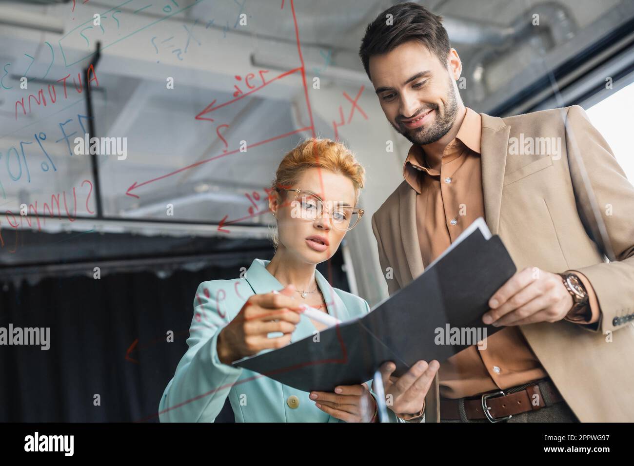 Eine Geschäftsfrau mit Brille, die sich Dokumente neben einem lächelnden Kollegen ansieht und eine Glaswand im Büro hat, ein Bild von der Börse Stockfoto