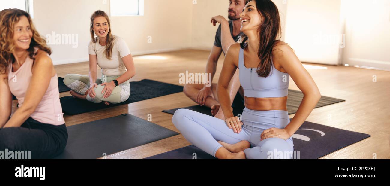 Glückliche Frauen, die zusammen lachen, während sie auf Trainingsmatten in einem Yoga-Studio sitzen. Fröhliche Frauen, die eine Pause von einer Hatha-Yoga-Session machen. Gruppe von Stockfoto