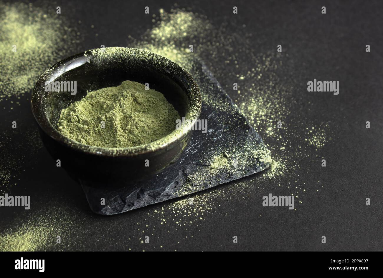 Grünes Pulver in einer kleinen Keramikschüssel, auf einem Steinständer, auf schwarzem Hintergrund. Der Hintergrund ist mit diesem Pulver bestreut. Super Food oder ma Stockfoto