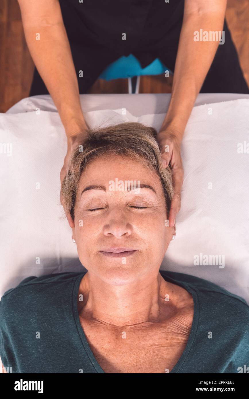 Von oben eine nicht erkennbare Therapeutin, die langsame Bewegungen mit den Fingern an den Kopf einer Seniorin anlegt, die mit geschlossenen Augen in der Klinik liegt Stockfoto