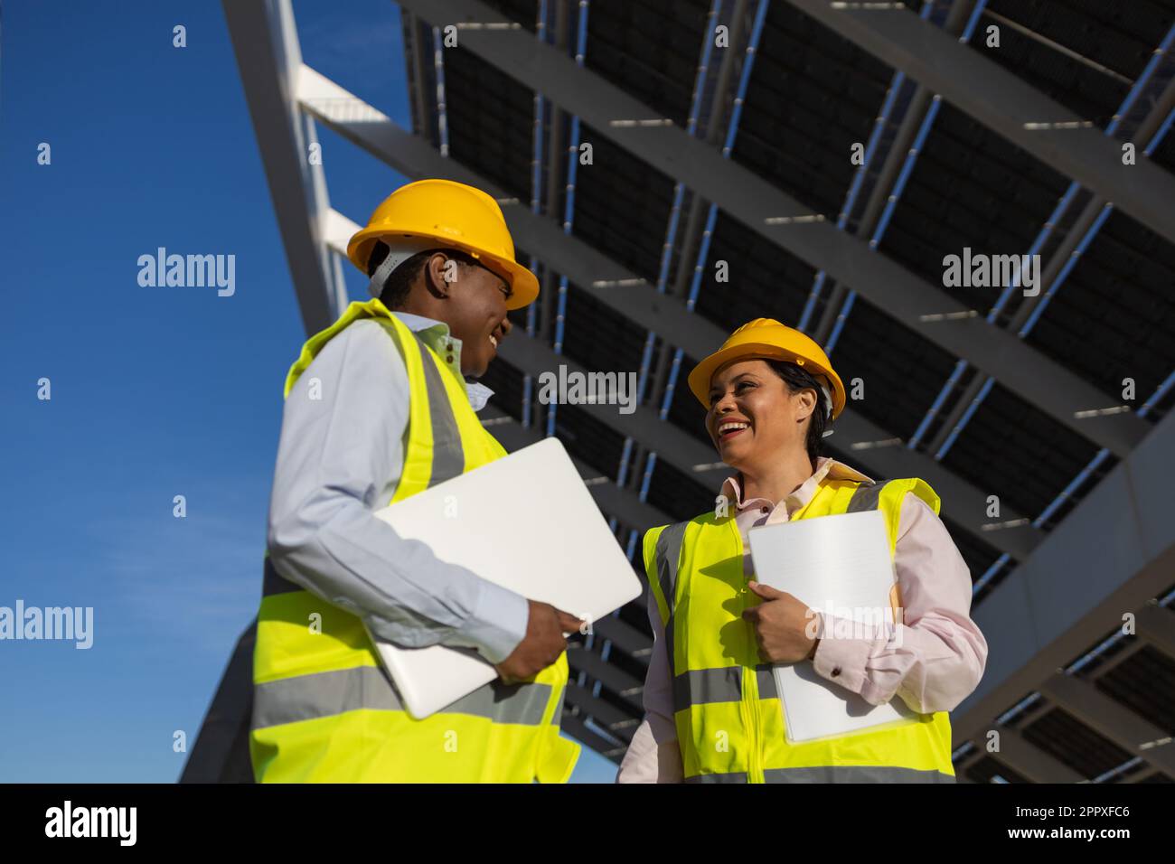 Kleiner Winkel junger schwarzer Ingenieurinnen in gelben Schutzhelmen und Westen, die Ordner mit Dokumenten und Laptop halten, während sie über nachhaltige Solarenergie sprechen Stockfoto