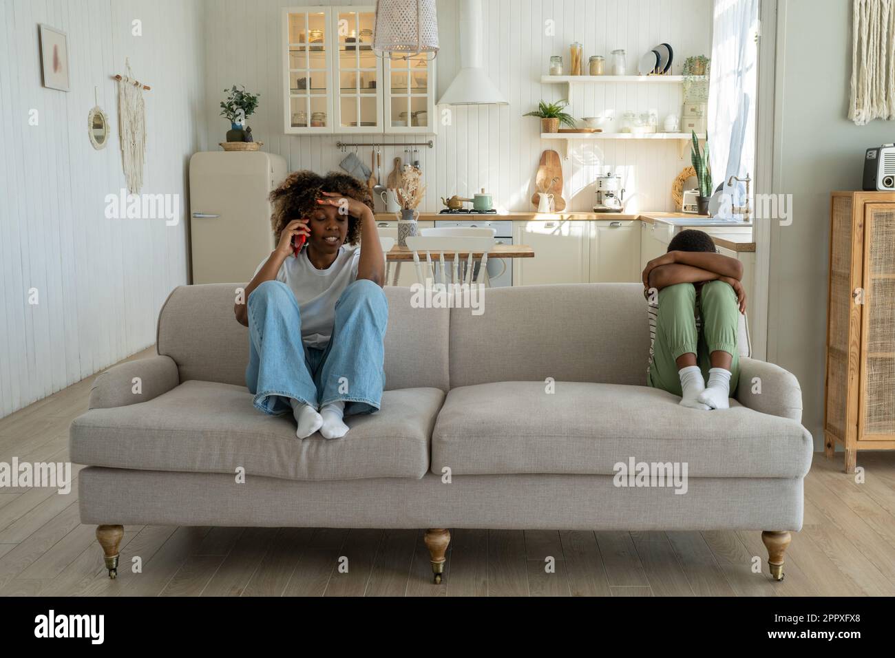 Verärgerte afroamerikanische Mutter, die mit einem beleidigten Sohn auf dem Sofa sitzt und telefoniert Stockfoto