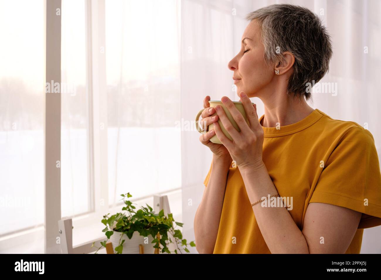 Seitenansicht einer gereiften Frau mit geschlossenen Augen in legerer Kleidung und leckerem frischen Heißgetränk in der Nähe des Fensters mit Vorhang in einem hellen, modernen Apartment Stockfoto