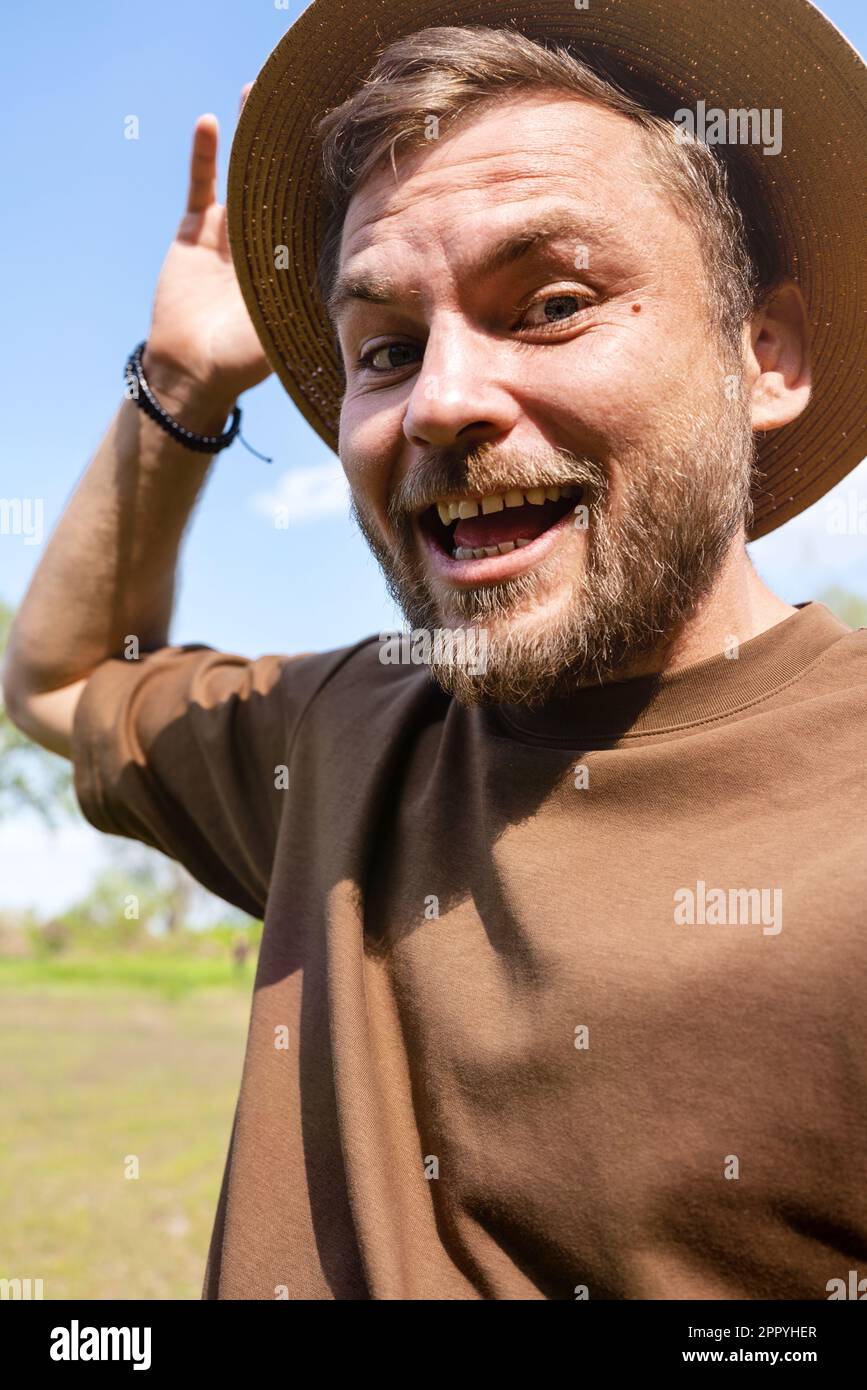 Ein junger erwachsener Mann mit Strohhut lächelt und macht ein Selfie, während er draußen spaziert. Stockfoto