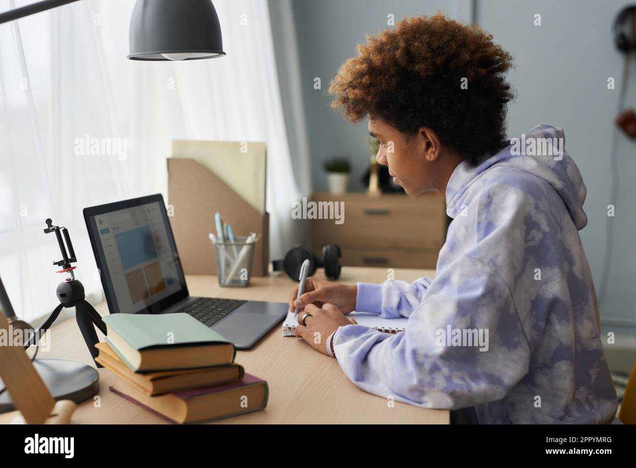 Seitenansicht eines jungen Schülers im Hoodie, der sich Notizen in einem Copybook macht und während der Schulaufgabe auf den Laptop-Bildschirm schaut Stockfoto