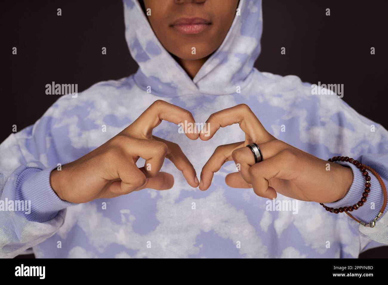 Nahaufnahme eines afroamerikanischen Teenagers in stilvollem Hoodie, der Herz mit seinen Fingern vor sich selbst macht, während er in Isolation im Studio posiert Stockfoto