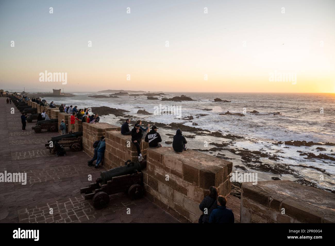 Gruppe von Personen, die auf den Stadtmauern des Hafens von Essaouira sitzen, um den Sonnenuntergang zu beobachten. Stockfoto