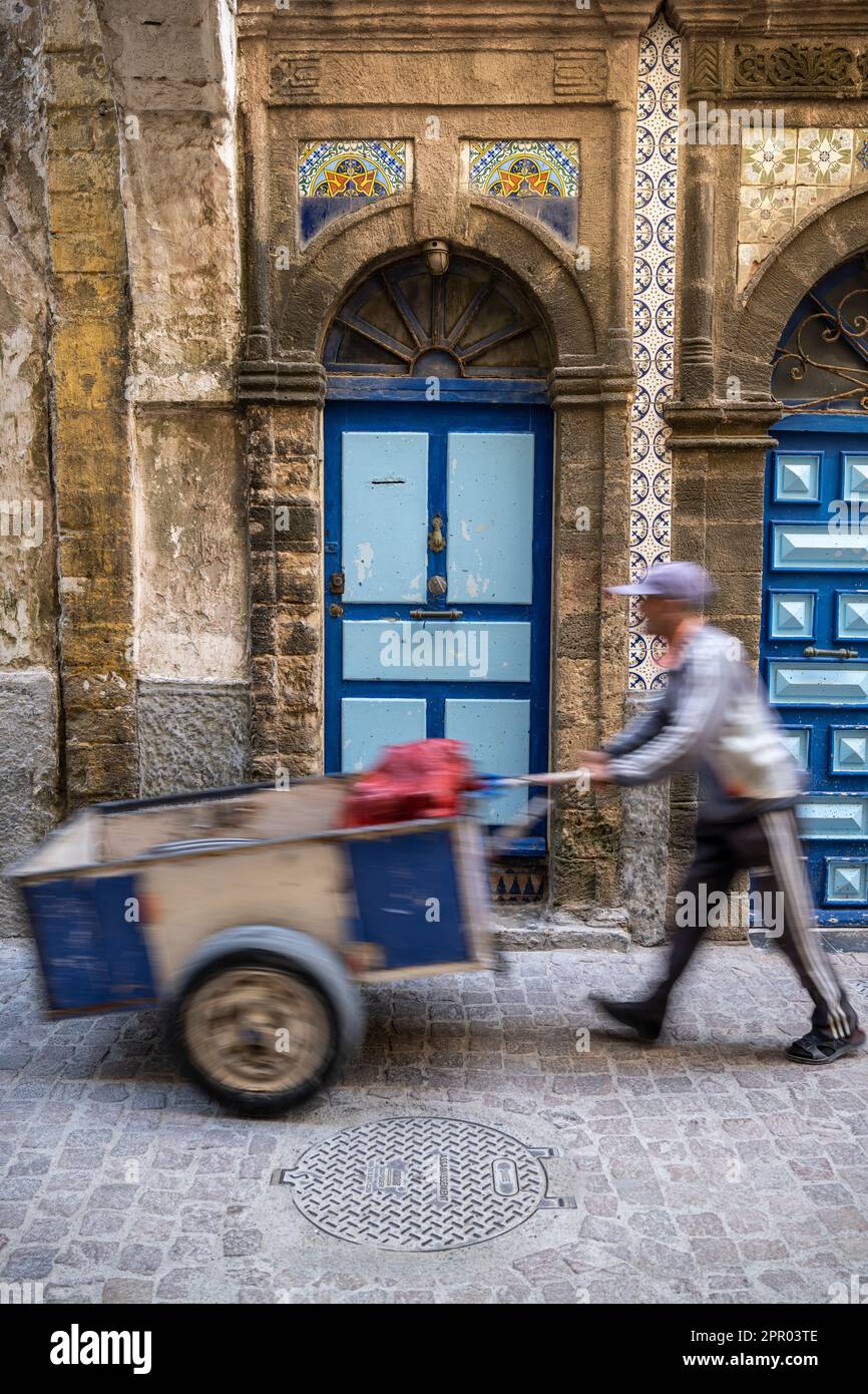 Silhouette eines Mannes, der einen Wagen vor die Eingangstür zu einem alten Haus in der Medina von Essaouira schiebt. Stockfoto