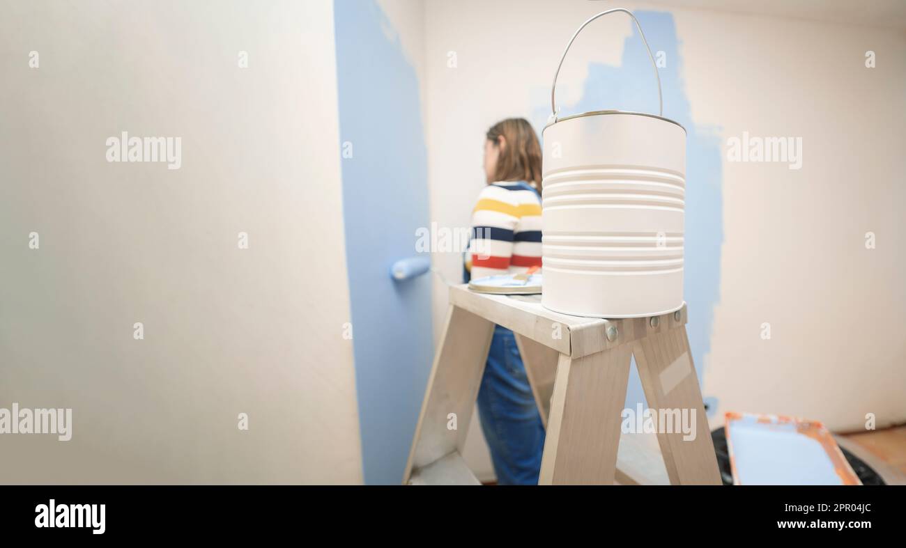 Nahaufnahme eines Glases Farbe auf einer Leiter mit dem Hintergrund einer stehenden Frau in Overalls und gestreifter Bluse, von hinten gesehen, die einen weißen Lack malt Stockfoto