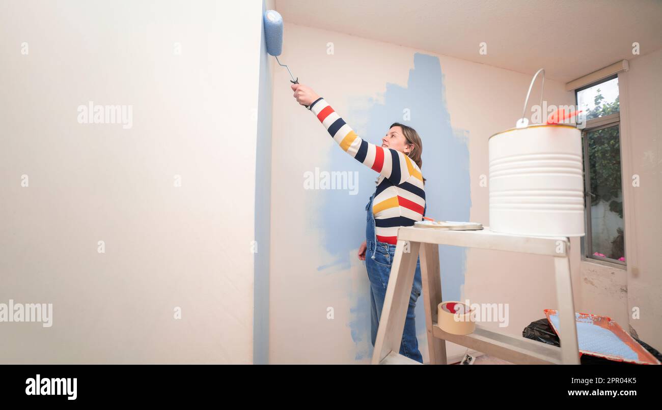 Nahaufnahme eines Glases Farbe auf einer Leiter mit dem Hintergrund einer stehenden Frau in Overalls und gestreifter Bluse, von hinten gesehen, die einen weißen Lack malt Stockfoto