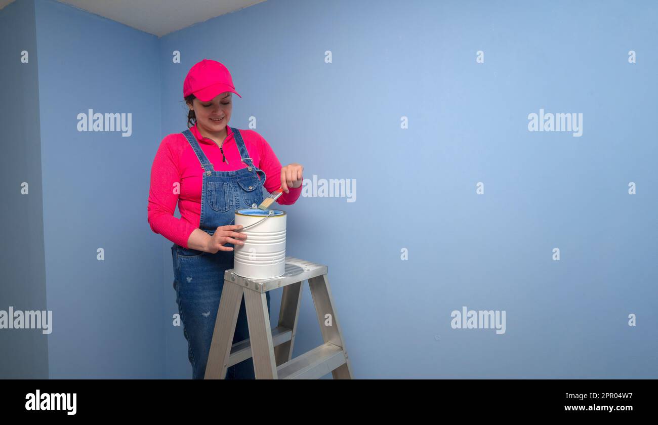 Wunderschöne Frau in Overalls und rotem T-Shirt, die auf einer Metallleiter steht und ihren Pinsel in ein Glas mit blauer Farbe neben einer blauen Wand taucht Stockfoto