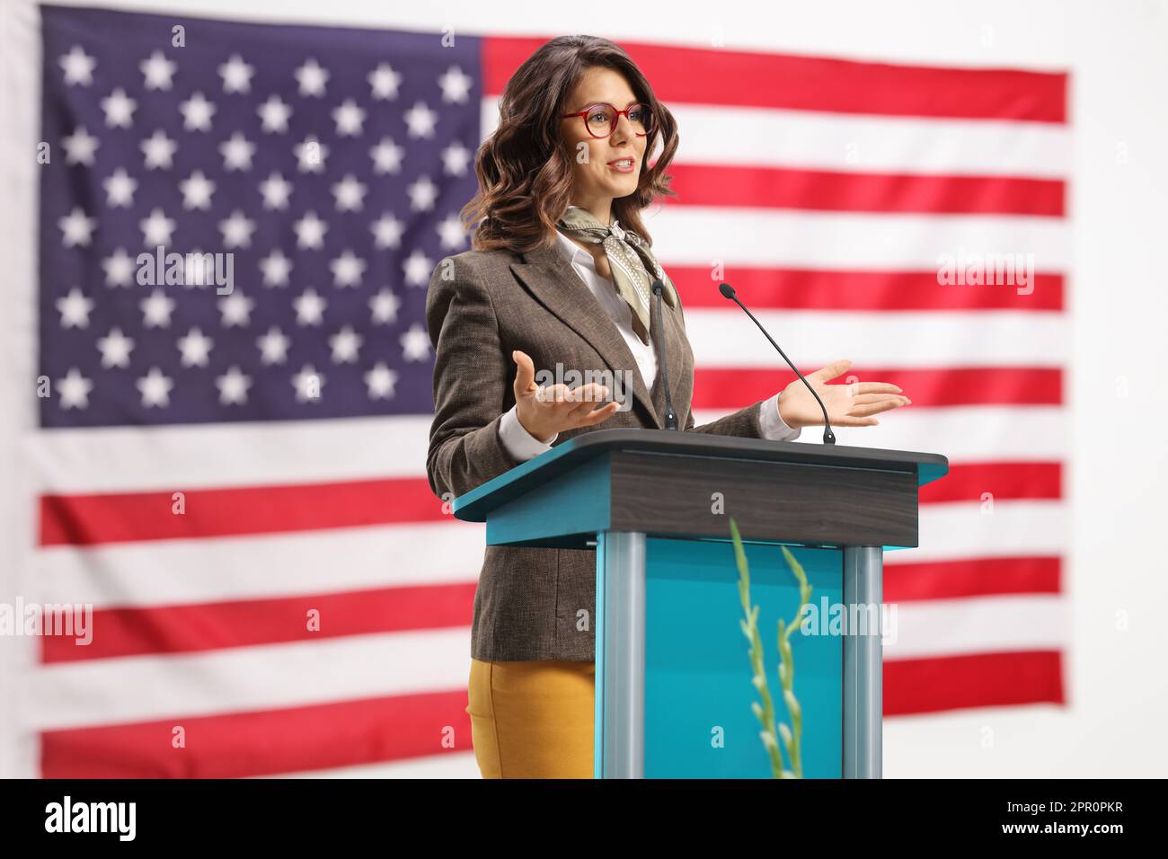 Junge Frau auf einem Podest, die eine Rede hält und Gesten mit Händen mit der US-Flagge im Hintergrund macht Stockfoto