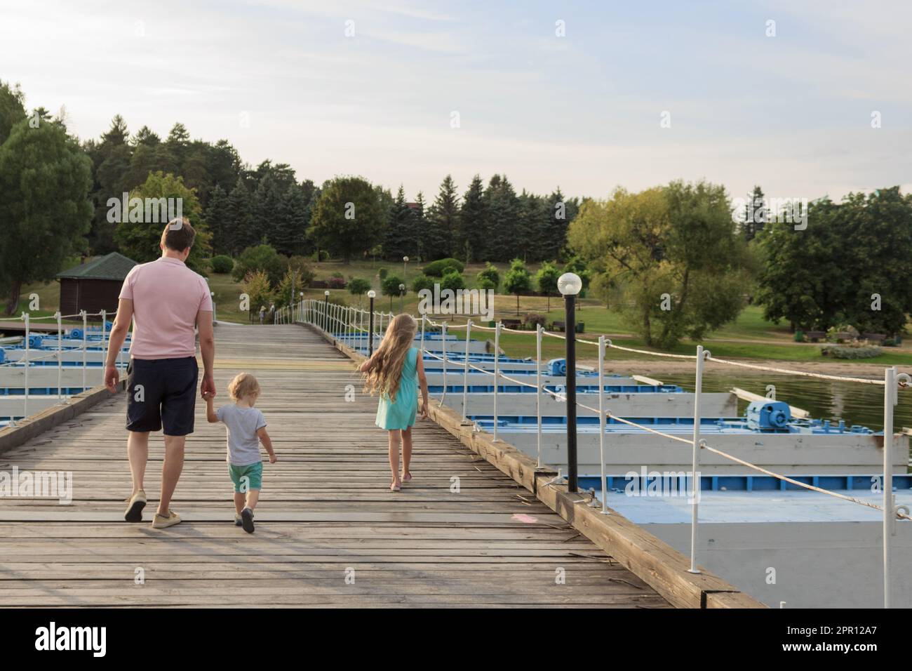 Zwei Generationen: Dad und zwei Kinder laufen am See im Park vorbei, Blick von hinten. Familie, Steg, Meer und Mann mit Kindern hinten. Stockfoto