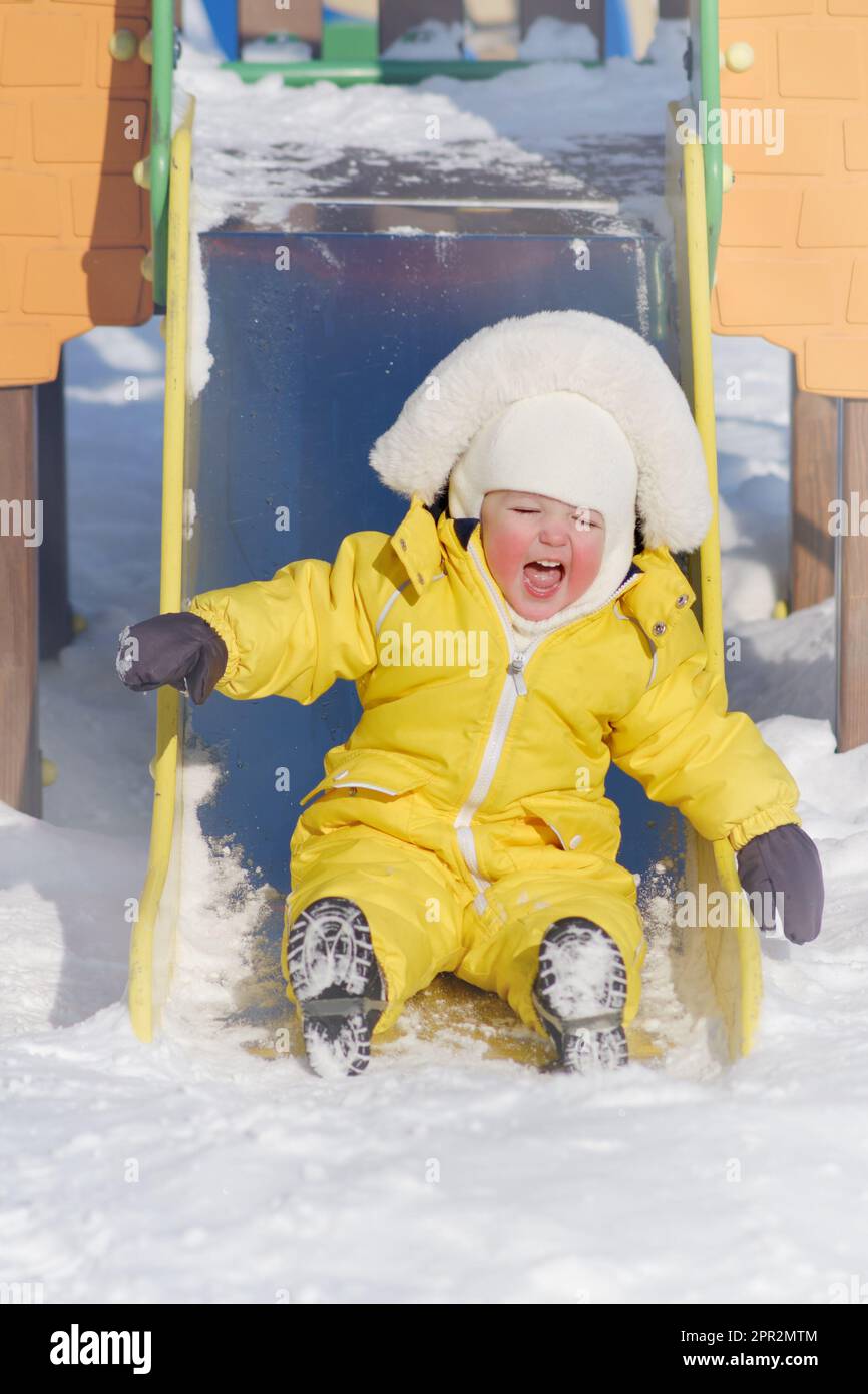 Der kleine Junge fährt auf einer Rutsche und spielt auf einem Winterspielplatz. Ein Kind in einem gelben Overall auf einer Kinderrutsche im Schnee. Ein Kind im Alter von einem Jahr Stockfoto