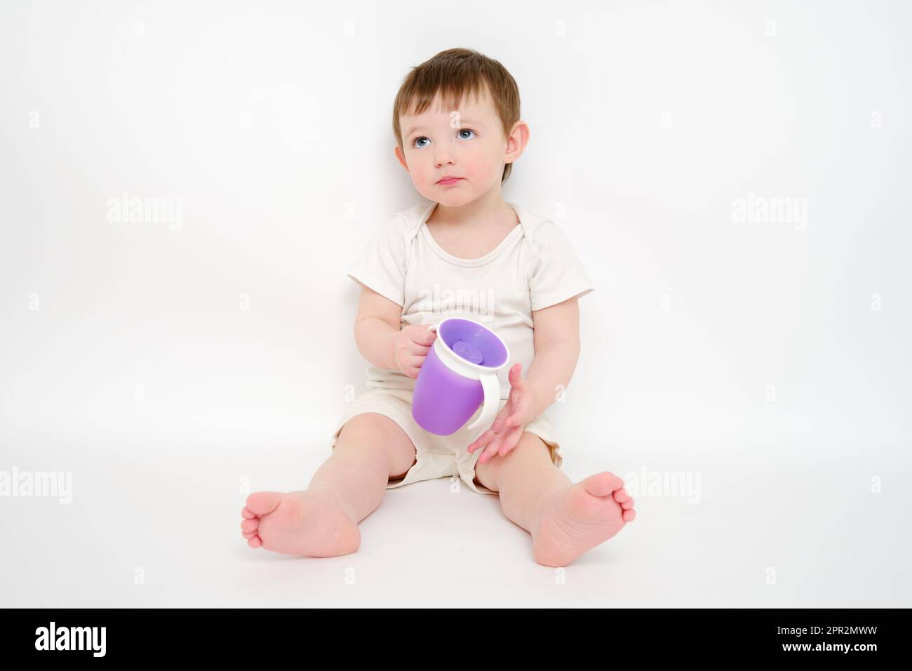 Fröhliches Baby trinkt Wasser aus einer Tasse auf weißem Studiohintergrund. Ruhendes Kind mit Tassensaft in den Händen. Kind etwa zwei Jahre alt (ein Jahr neun Monate) Stockfoto