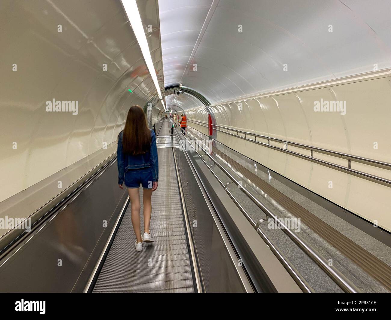 Ein hübsches Mädchen geht am Travolator im U-Bahn-Tunnel entlang, um zu einer anderen U-Bahn-Station zu gehen. Blick von hinten. Stockfoto
