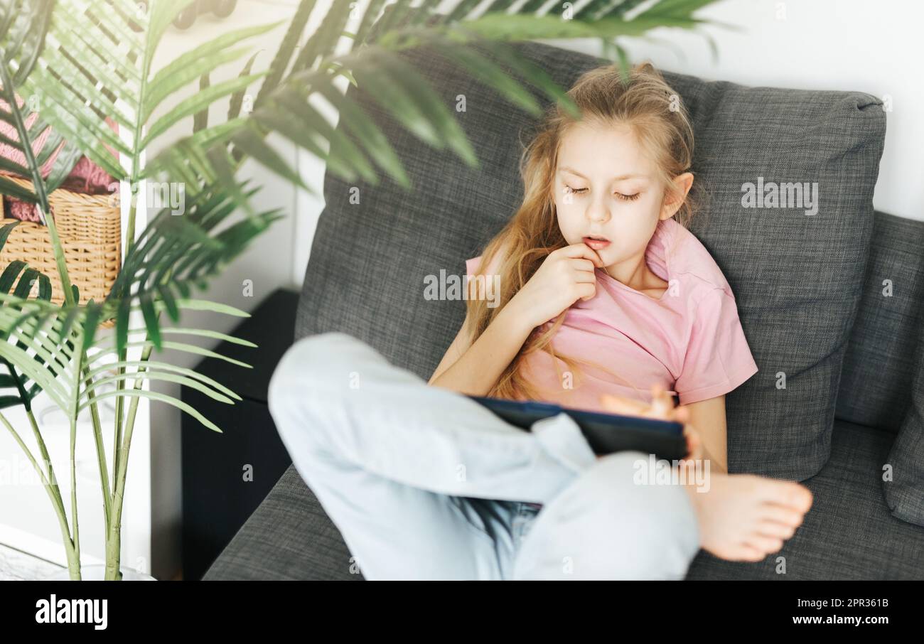 Ein kleines Mädchen, das auf der Couch liegt, mit einem elektronischen Tablet. Einsatz von drahtlosen Geräten für den Bildungsbereich. Stockfoto