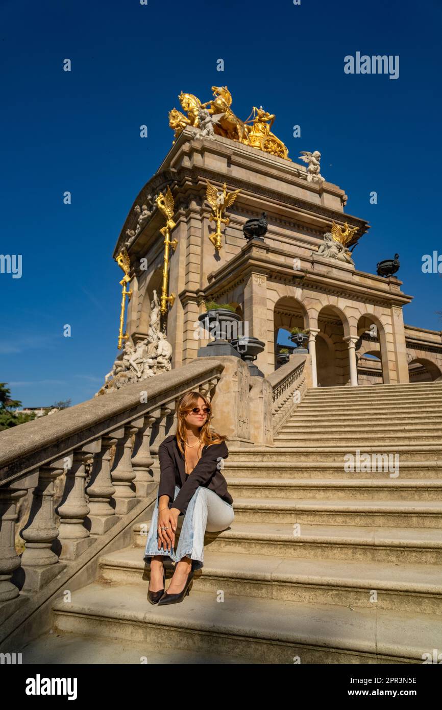 Junge, gebräunte, schöne Frau mit geschlossenen Augen in einem schwarzen Oberteil und Jeans mit Sonnenbrille und langen Haaren, die stilvoll und elegant in der Sonne sitzt Stockfoto