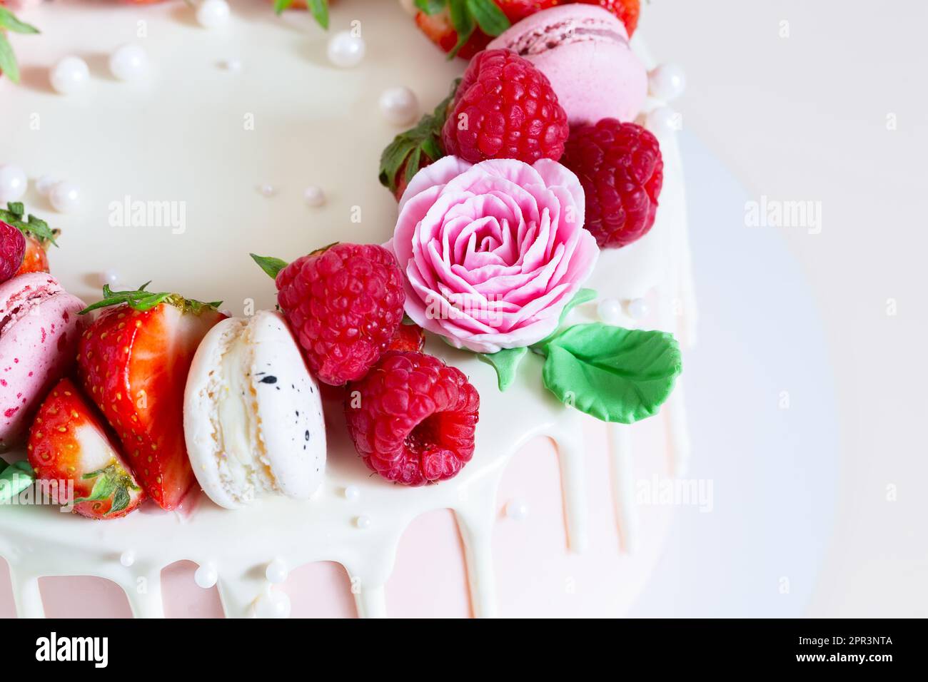 Süßer Geburtstagskuchen mit Beeren, Makronen und Blumendekor auf dem Tisch. Wunderschöner rosa Kuchen dekoriert mit Makronen, Himbeeren, Erdbeeren und Zucker Stockfoto