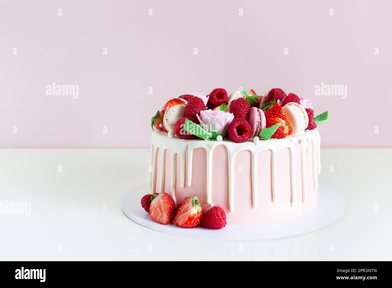 Süßer Geburtstagskuchen mit Beeren, Makronen und Blumendekor auf dem Tisch. Wunderschöner rosa Kuchen dekoriert mit Makronen, Himbeeren, Erdbeeren und Zucker Stockfoto