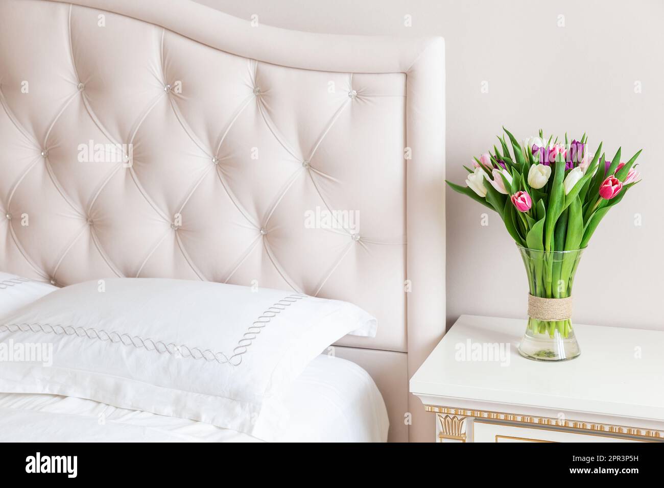 Schlafzimmer in sanften hellen Farben. Vase mit bunten Tulpen in hellem, gemütlichen Schlafzimmer-Interieur. Wohneinrichtung mit rosa Tulpen in einer Vase auf einer hellen Schlafzimmerrückseite Stockfoto