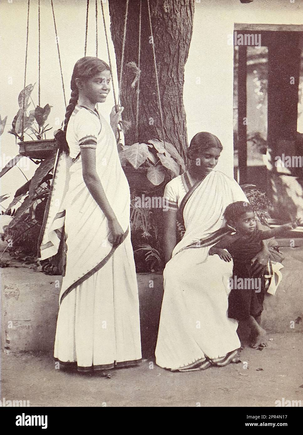 Ältere indische Mädchen mit einer jüngeren, neben einem Baum mit Schaukeln und einer Topfpflanze. In der Dohnavur-Anlage. Halbtongravierung auf einem Foto von Mr. Penn aus Ootacamund (jetzt bekannt als OOTO), in der Region Tamil Nadu in Südindien, c1912. Das Bild bezieht sich auf das Kinderzimmer und das Gelände der Church of England Zenana Mission in Dohnavur, ebenfalls in Tamil Nadu und etwa 30 Meilen von der Südspitze Indiens entfernt. Das Gelände wurde von Amy Wilson-Carmichael betrieben und versuchte, das Christentum bei Babys und Kindern zu fördern (damals nur Mädchen, von denen viele Opfer der Tempelprostitution waren). Stockfoto