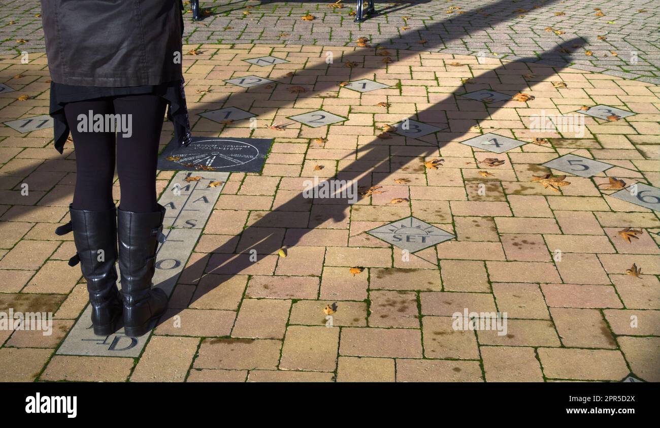 Person, die mit dem Human Millenium Sun Dial steht, das auf dem Bürgersteig, Street, Ringwood England UK eingebaut ist Stockfoto