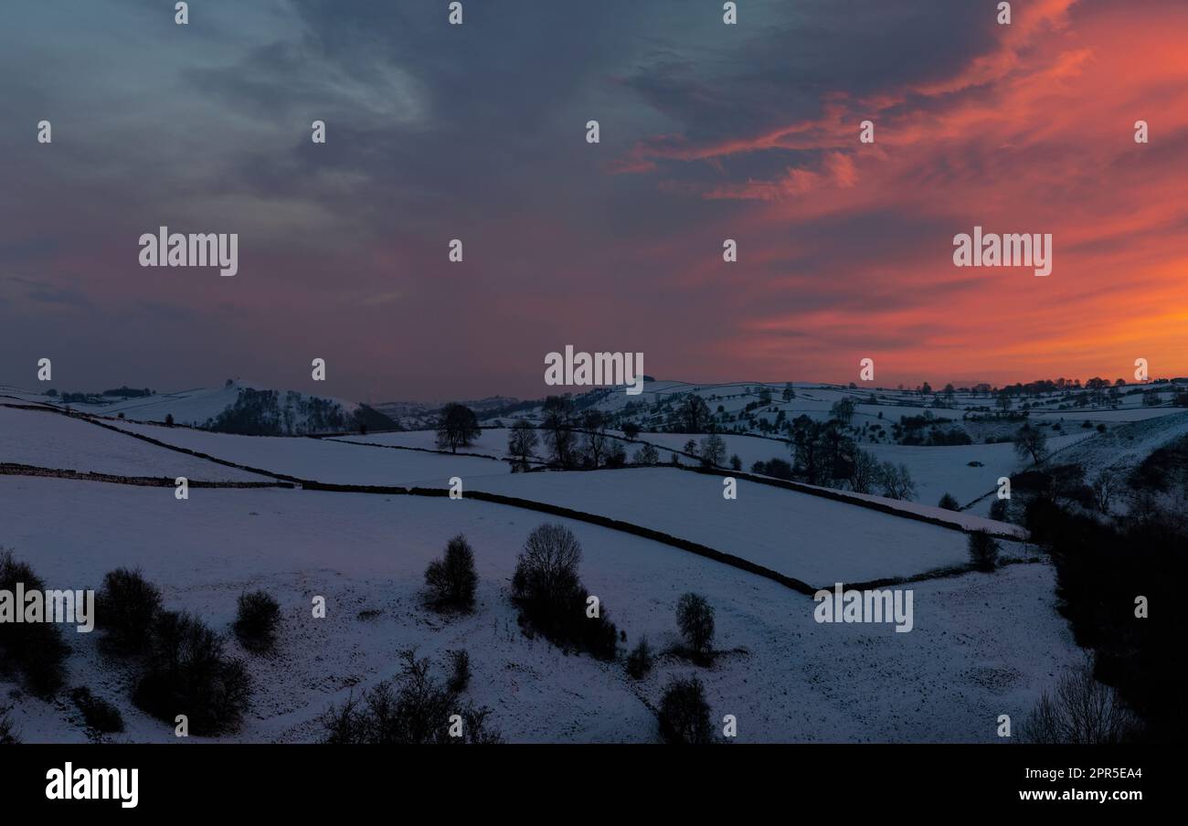 Die schneebedeckte Landschaft von Staffordshire im Peak District, kurz nach Sonnenuntergang mit noch etwas Rot am Himmel. Ackerland, Bäume und Trockenmauern Stockfoto