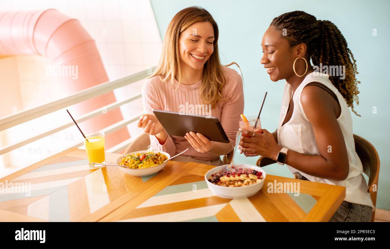 Zwei hübsche junge schwarze und weiße Frau, die sich amüsiert, frische Säfte trinkt und ein gesundes Frühstück im Café hat Stockfoto
