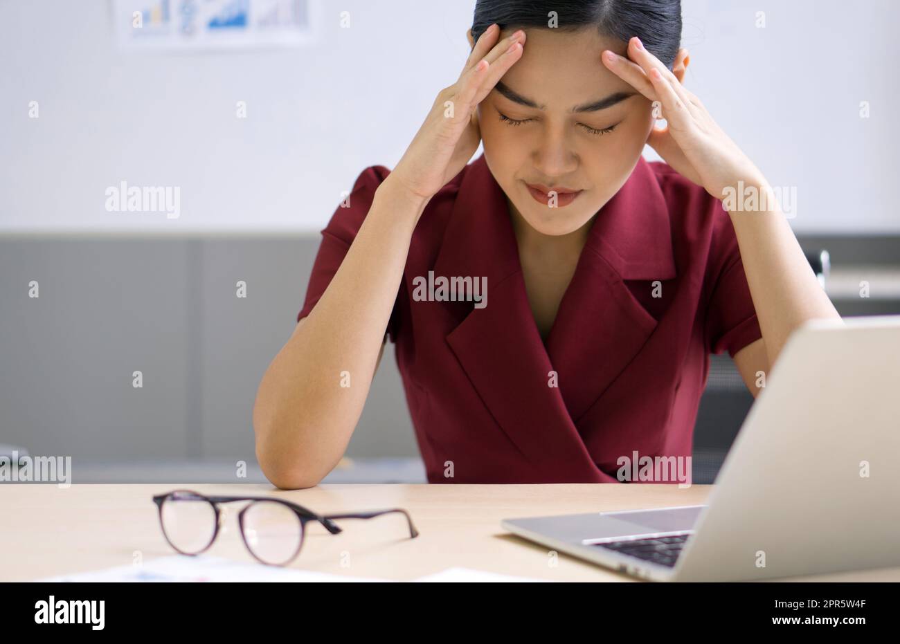 Die junge asiatische Geschäftsfrau hält ihren Tempel mit beiden Händen und sitzt im Büro. Das Konzept von Arbeitslosen, Traurigkeit, Depressionen und menschlichen Problemen. Stockfoto