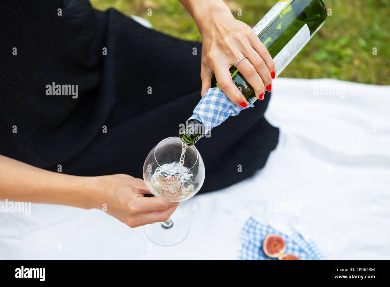 Romantisches Picknick im Park auf dem Gras, köstliches Essen: Korb, Wein, Trauben, Feigen, Käse, blaue karierte Tischdecke, zwei Gläser Wein. Mädchen schüttet Wein in ein Glas. Outdoor-Freizeitkonzept. Stockfoto