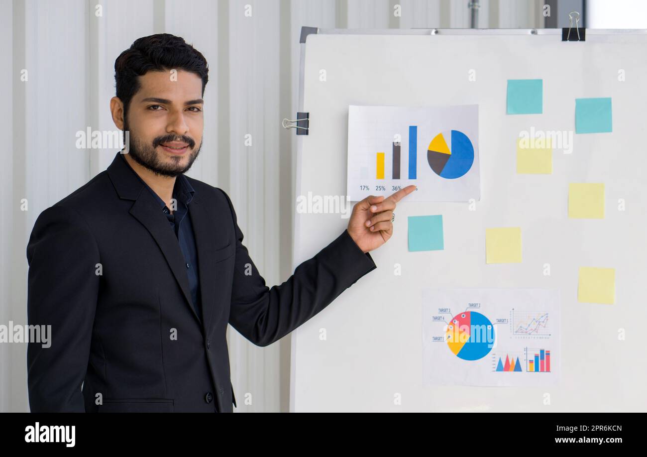 Junger, gewellter Geschäftsmann mit Schnurrbart und Bart in schwarzem Anzug, der mit dem Finger auf die Tortenkarte auf dem Whiteboard zeigt. Aus- und Weiterbildungskonzepte. Stockfoto