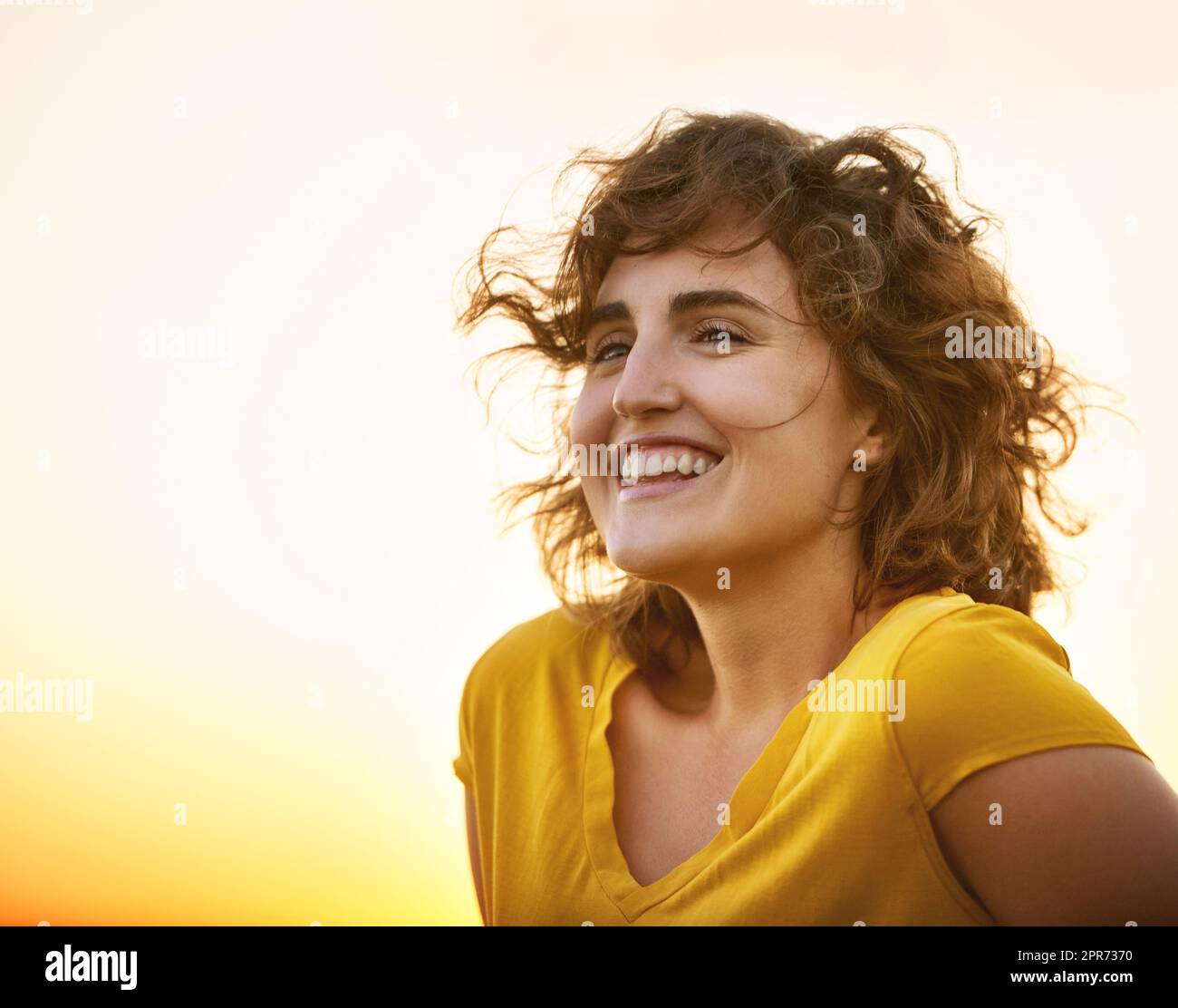 Shes hatte Sonnenschein in ihrer Seele. Aufnahme einer fröhlichen jungen Frau, die lächelt, während sie beim Sonnenuntergang draußen steht. Stockfoto