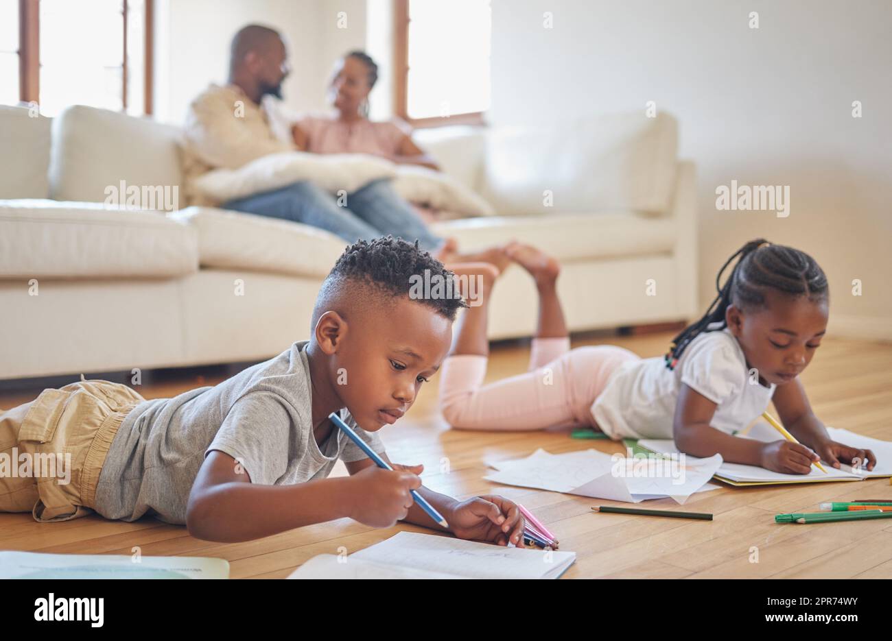 Kleine Jungen und Mädchen malen mit Malstiften auf dem Wohnzimmerboden, während sich ihre Eltern auf dem Sofa entspannen. Kleine Kinder, Schwestern und Geschwister, die sich in der Familienzeit zuhause einfärben Stockfoto