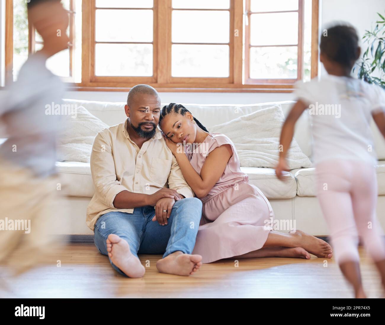 Ein müdes afroamerikanisches Paar, das auf dem Loungeboden saß und müde aussah, während ihre Hyperkinder um sie herum spielten. Zwei schwarze Geschwister, die Spaß haben, während ihre erschöpften Eltern sich entspannen Stockfoto