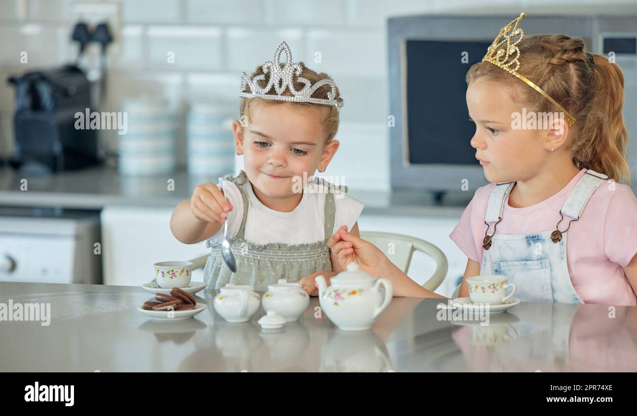 Zwei kleine Mädchen, die zu Hause eine Teeparty machen. Geschwisterfreunde tragen Tiaras, während sie mit dem Teeset spielen und Kekse am Küchentisch trinken. Sis Stockfoto
