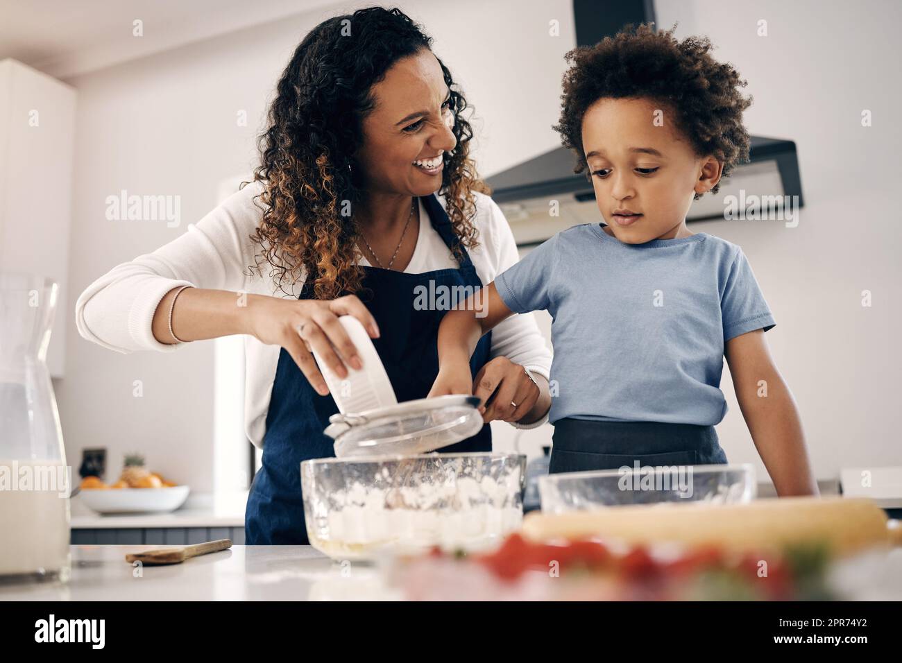 Ein süßer kleiner Junge mit Afro-Backen in der Küche zu Hause mit seiner mutter. Fröhliche Mischrasse Frau, die mit Hilfe ihres Sohnes Mehl sieben und Zutaten mischen kann. Das Backen ist eine Bindungsaktivität Stockfoto