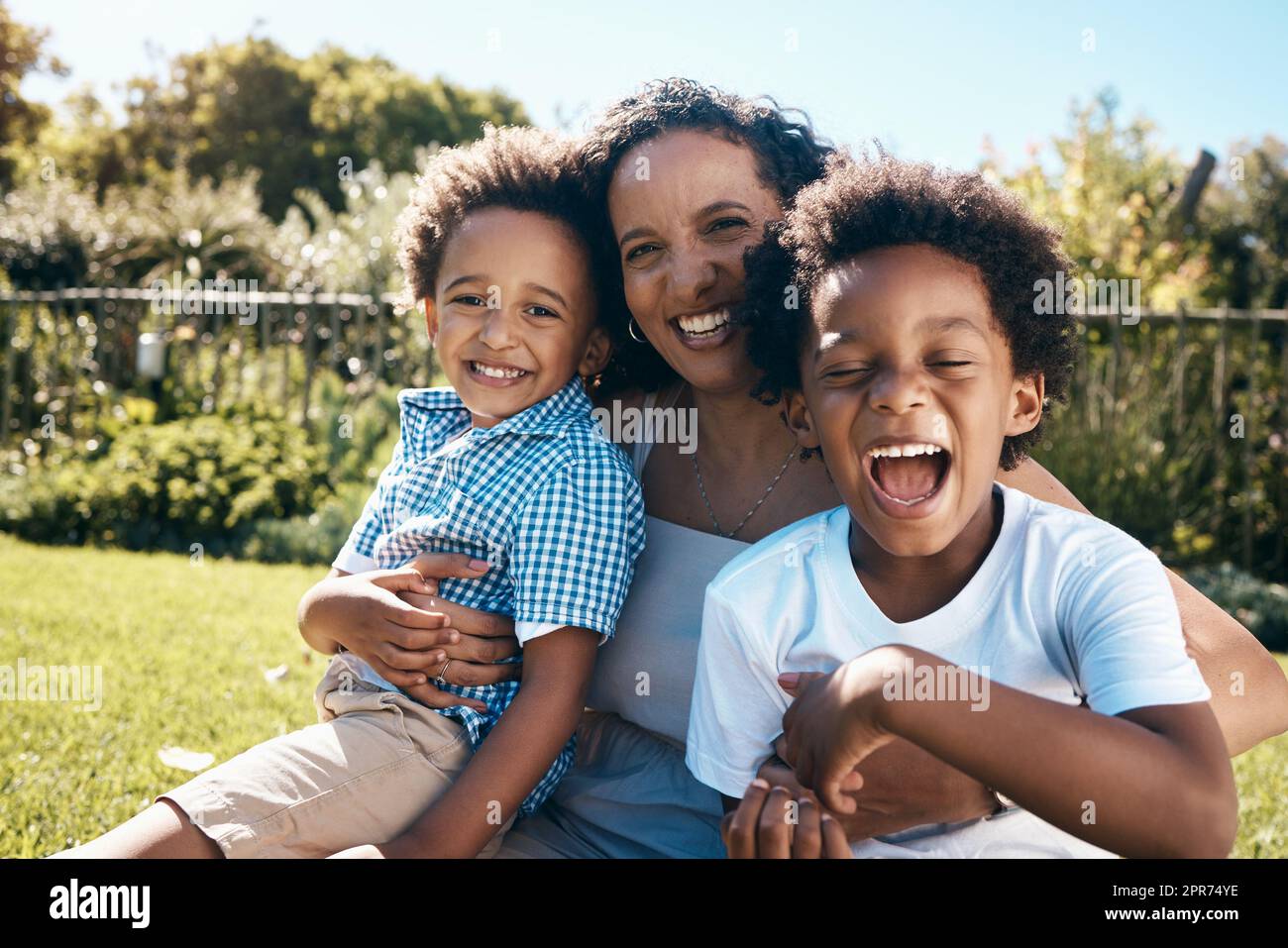 Aufgeregter kleiner Junge mit Afro sitzt draußen auf dem Gras mit seiner Mutter und seinem Bruder. Eine energiegeladene afroamerikanische Familie verbringt Zeit im Park oder im Garten. Mom umarmt zwei Söhne Stockfoto