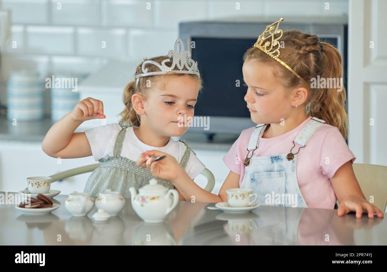 Zwei kleine Mädchen feiern zu Hause eine Prinzessinnen-Teeparty. Geschwister oder Freunde, die beim Spielen mit dem Teeset Diademe tragen und Kekse am Küchentisch essen. Schwestern verstehen sich gut und spielen zusammen Stockfoto
