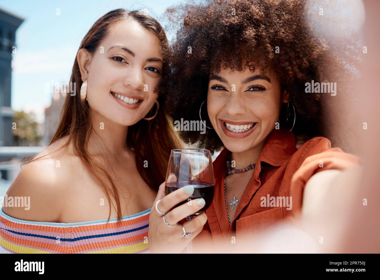 Zwei wunderschöne Mischwettrennen lächelnde Frauen machen ein Selfie, während sie draußen auf einer Party ein Glas Rotwein genießen. Hispanische Frau mit coolem Afro-Haar, die an einem sonnigen Tag ihre Zeit mit einer Freundin genießt und dabei ein Getränk genießt Stockfoto