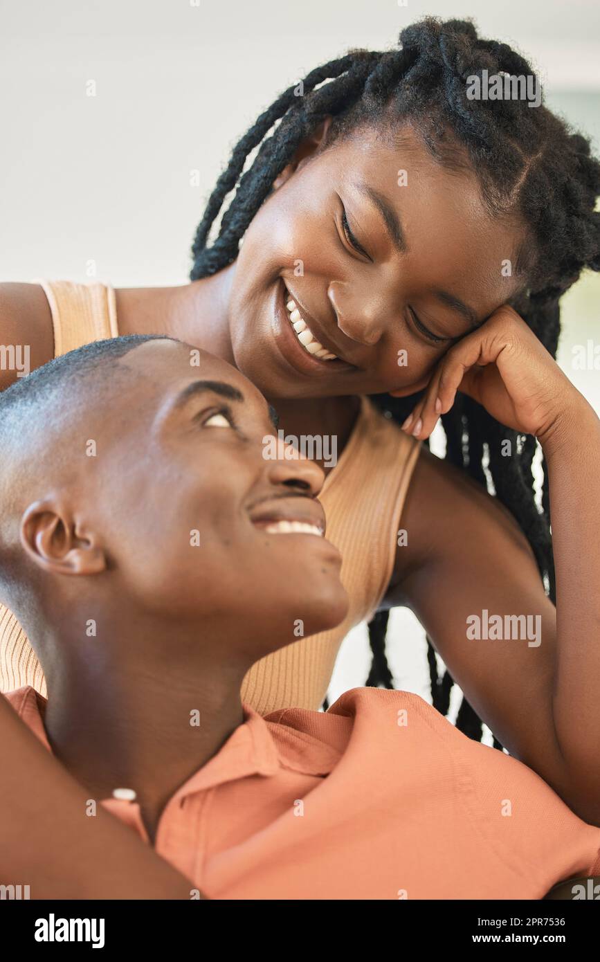 Ein junges, fröhliches afroamerikanisches Paar, das sich zu Hause anfreundet und eine entspannte Zeit zusammen genießt. Eine schwarze Frau, die lächelt, während sie sich umarmt und ihren Freund ansieht, der zusammensitzt und redet Stockfoto