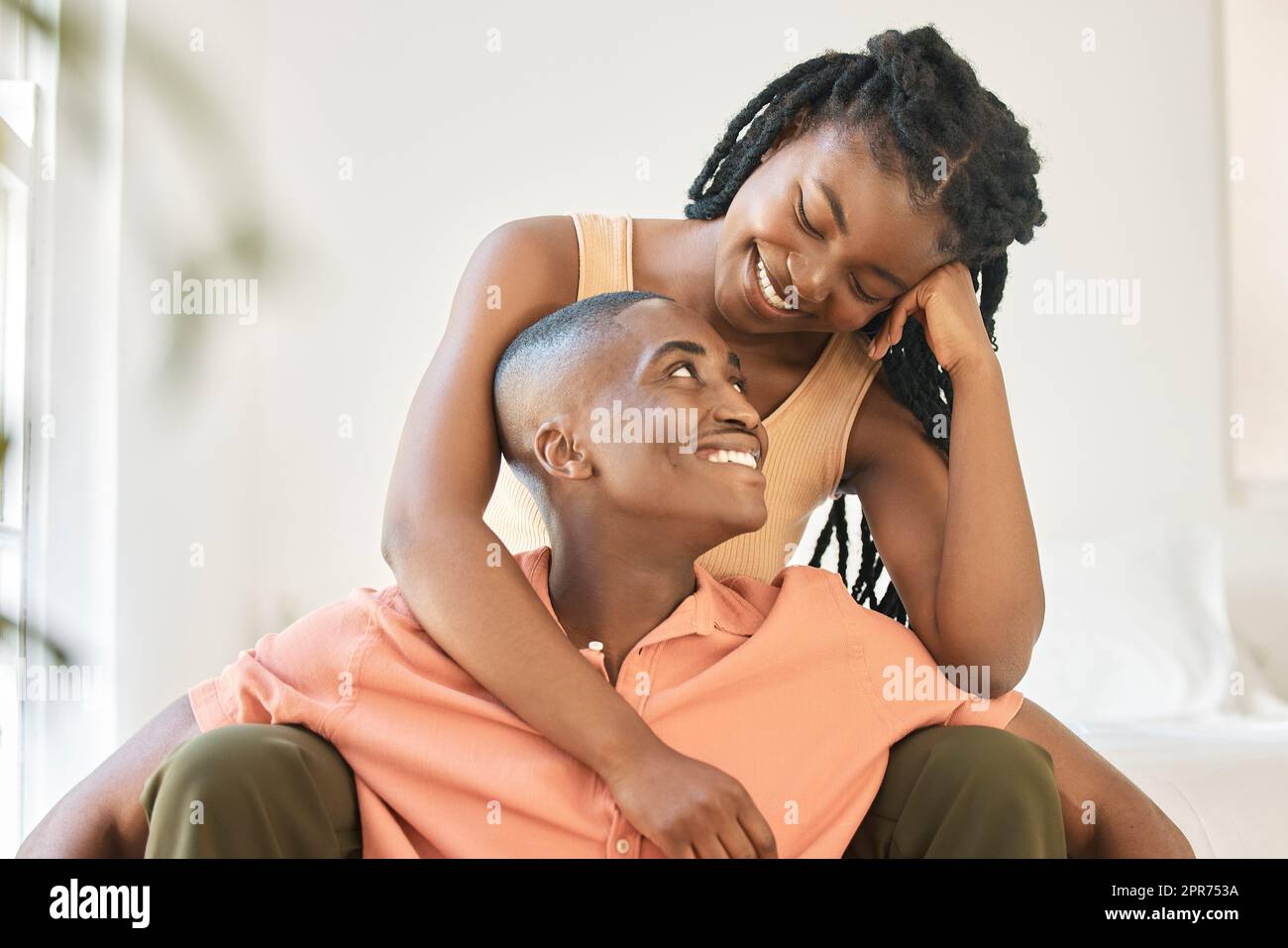 Ein junges, glückliches, sorgenfreies und fröhliches afroamerikanisches Paar, das sich zu Hause anfreundet und eine entspannte Zeit zusammen genießt. Eine schwarze Frau, die lächelt, während sie sich umarmt und ihren Freund ansieht, der zusammen sitzt Stockfoto
